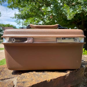 May include: A brown hardshell train case with a textured exterior finish. The case features a top handle, a silver-toned metal trim along the center, and two chrome-plated locking mechanisms with keyholes positioned on the front panel.