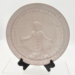 May include: A circular light gray ceramic plate featuring a raised relief of a figure with a halo, positioned in front of a cross. The text 19-KING OF KINGS-70 is embossed along the bottom edge, and a small star is visible in the upper right corner.
