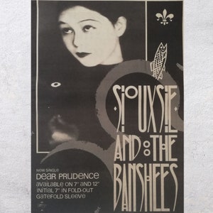 May include: Black and white promotional poster for the band Siouxsie and the Banshees. The poster features a black and white photo of Siouxsie Sioux, the band's lead singer, and the band's name in a stylized font. The text "NEW SINGLE DEAR PRUDENCE AVAILABLE ON 7" AND 12" INITIAL 7" IN FOLD-OUT GATEFOLD SLEEVE" is printed at the bottom of the poster.