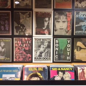 Puede incluir: Un collage de pared de p&oacute;sters de m&uacute;sica vintage con artistas como Bowie, The Rolling Stones, Tom Waits, Nick Cave, U2 y Sonic Youth. Los p&oacute;sters est&aacute;n enmarcados en marcos negros y blancos y se exhiben en una pared con un fondo amarillo.