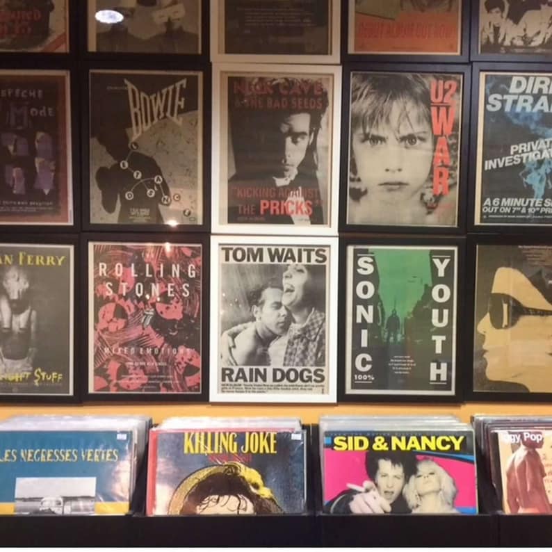 Puede incluir: Un collage de pared de p&oacute;sters de m&uacute;sica vintage con artistas como Bowie, The Rolling Stones, Tom Waits, Nick Cave, U2 y Sonic Youth. Los p&oacute;sters est&aacute;n enmarcados en marcos negros y blancos y se exhiben en una pared con un fondo amarillo.