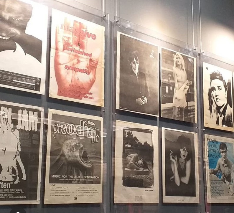 May include: A wall display of framed posters featuring various music artists and bands, including a poster with the text "Massive Unfinished Sympathy" and a poster with the text "Prodigy Music for the Jilted Generation".