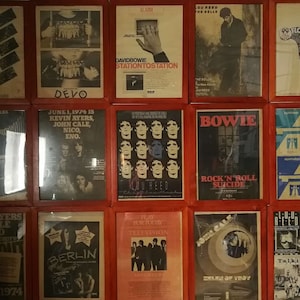 Puede incluir: Un collage de pared de carteles enmarcados con varios m&uacute;sicos y bandas, incluyendo Devo, David Bowie, Lou Reed, The Bells y John Cale. Los carteles est&aacute;n todos en marcos rojos y tienen una est&eacute;tica vintage.