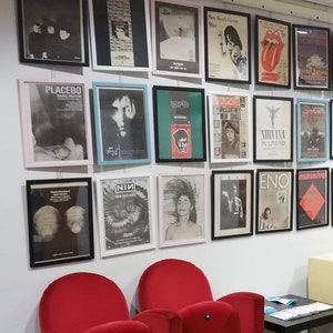 Puede incluir: Una pared con una colecci&oacute;n de p&oacute;sters de m&uacute;sica enmarcados que presentan bandas como Placebo, Nirvana y NIN. Los p&oacute;sters son de varios colores y estilos, y algunos incluyen t&iacute;tulos de &aacute;lbumes y logotipos de bandas.