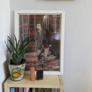 Puede incluir: Un p&oacute;ster enmarcado en blanco con el texto "IN SEARCH OF STOUP" y una foto en blanco y negro de un hombre. El p&oacute;ster est&aacute; colgado en una pared sobre un estante con libros y bloques de madera que deletrean "IB".