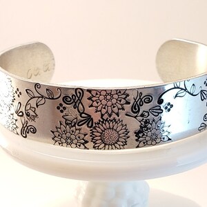 May include: Silver cuff bracelet with a black floral design etched into the metal.