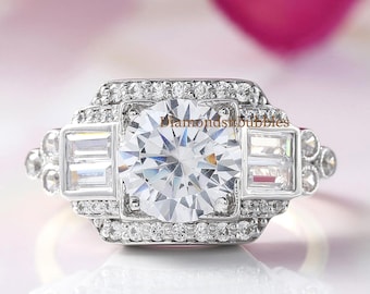 May include: A silver engagement ring with a large, round, clear center stone and smaller, rectangular side stones. The ring is surrounded by smaller accent stones. The background features blurred pink flower petals.