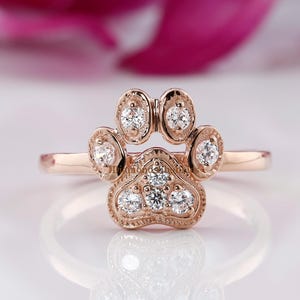 Op de afbeelding: Rosegouden ring in de vorm van een pootafdruk, bezet met heldere edelstenen. De ring heeft een pootafdrukontwerp met vijf ronde edelstenen die in het midden en aan de uiteinden van de voetzolen zijn gezet. De ring heeft een dunne band.