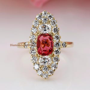 May include: A gold ring featuring a central oval-cut, reddish-pink gemstone, surrounded by a halo of smaller, clear gemstones. The ring has a detailed, elongated design with additional small gemstones on the band. The ring is set against a soft, blurred background.