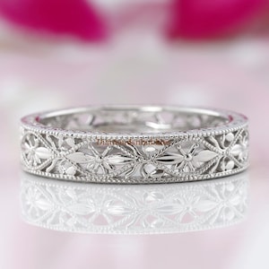 以下が含まれることがあります： 精巧な花柄が施されたシルバーリング。リングは、花と葉の繰り返し模様で、繊細で詳細な外観を作り出しています。リングは、柔らかくぼやけた背景に配置されています。