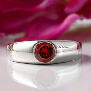 以下が含まれることがあります： ベゼルセッティングに赤い宝石がセットされたシルバーリング。リングは白い表面の上に置かれています。