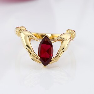Peut inclure: Une bague dorée ornée d'une pierre rouge foncé taillée en marquise. La conception de la bague incorpore deux mains stylisées tenant la pierre précieuse. La bague est posée sur un fond blanc, avec le texte "Diamondenbubbles" visible.