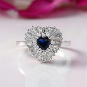 Puede incluir: Un anillo de plata en forma de corazón con un gran zafiro azul rodeado de pequeños diamantes blancos. El anillo está engastado con diamantes de forma baguette alrededor del zafiro.