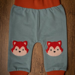 May include: Baby trousers in a soft teal fabric with orange trim at the waist and ankles. Two embroidered fox faces are on the knees. The trousers are made of a comfortable material and are suitable for infants and toddlers.