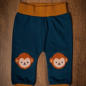 May include: Teal baby trousers with a mustard yellow waistband and cuffs. The trousers feature two embroidered monkey faces on the knees. The background is a wooden surface.