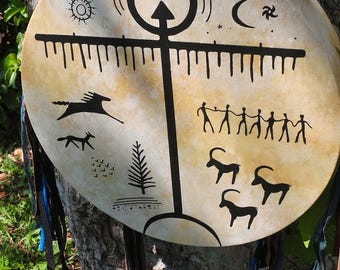 May include: A hand-painted Native American drum with a black and white design. The drum features a central cross with a circle at the top, surrounded by various symbols including a sun, moon, stars, and animals. The drum is decorated with blue and brown leather fringe.