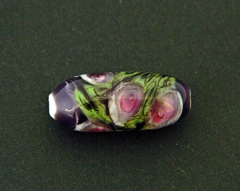 May include: An oblong glass bead with a floral design. The bead features a base of deep purple and white at the ends, with a central design of pink, green, and white swirls. The bead is approximately 2.5 cm long.