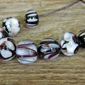 Könnte beinhalten: Eine Sammlung von Glasperlen in verschiedenen Designs. Einige Perlen sind klar mit weißen und schwarzen Mustern, andere weisen Wirbel aus Lila, Weiß und Silber auf. Die Perlen sind auf einer braunen Schnur aufgereiht und zeigen ihre einzigartigen Texturen und Farben.
