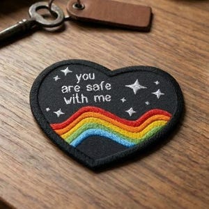 May include: A black heart-shaped embroidered patch with the text "you are safe with me" and a rainbow design. White stars surround the patch. A key, notebook, and candle are also on the wooden surface.