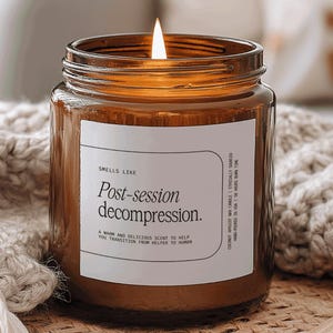May include: A lit amber glass jar candle with a white label that reads "Post-session decompression." The candle sits on a woven surface next to a beige knitted blanket. The candle's flame is lit, and the overall aesthetic is cozy and inviting.