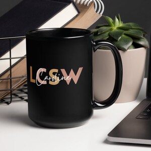 以下が含まれることがあります： 白い文字で「LCSW Caroline」とスタイリッシュなフォントで書かれた黒いセラミック製のコーヒーカップ。 「LCSW」の文字は、ピーチとライトブラウンの色です。