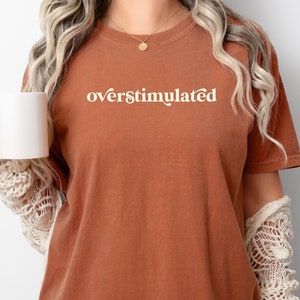 Può includere: Una donna indossa una maglietta color ruggine con la scritta "overstimulated" stampata in bianco. Sta tenendo una tazza bianca nella sua mano sinistra.