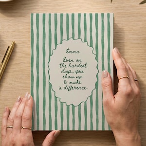 Könnte beinhalten: Ein Notizbuch mit grün gestreiftem Einband und dem Text "Emma Even on the hardest days, you show up & make a difference." Ein goldener Stift, Klebeband, Büroklammern und eine kleine Vase mit Grün sind ebenfalls zu sehen.