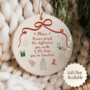 Puede incluir: Un adorno redondo de cerámica blanca con un gráfico de cinta roja en la parte superior y el texto: Olivia, Never forget the difference you make & the lives you've touched. Incluye pequeñas ilustraciones verdes y rojas de un regalo, un árbol y una taza.