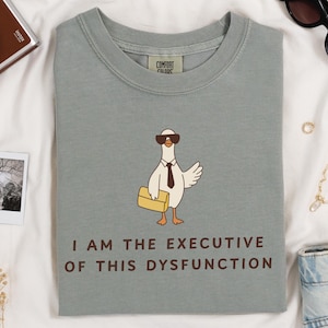 Puede incluir: Camiseta verde salvia con un dibujo de un ganso con gafas de sol y corbata, sosteniendo un maletín. El texto "I AM THE EXECUTIVE OF THIS DYSFUNCTION" está impreso debajo. La camiseta está doblada sobre una superficie blanca.