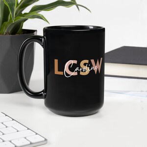 以下が含まれることがあります： 白いデザインで「LCSW Caroline」とカラフルな文字で書かれた黒いセラミックマグカップ