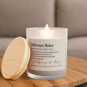 May include: A lit candle in a frosted glass jar with a wooden lid. The label reads "Difference Maker" with a definition and the phrase "for the difference you make - thank you." The candle is on a wooden table.