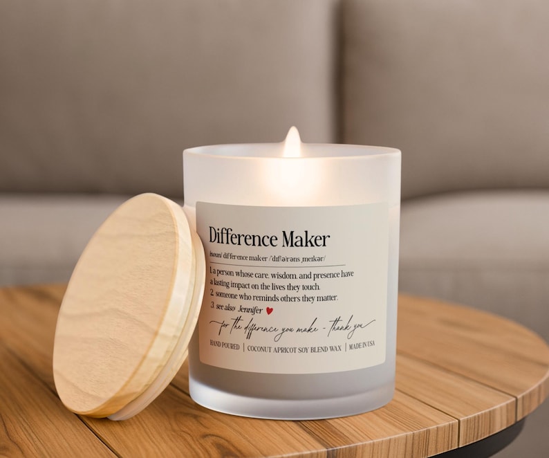 May include: A lit candle in a frosted glass jar with a wooden lid. The label reads "Difference Maker" with a definition and the phrase "for the difference you make - thank you." The candle is on a wooden table.