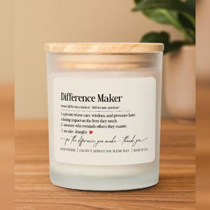 May include: A frosted glass candle with a wooden lid. The label reads "Difference Maker" with a definition and a handwritten message. The candle is hand-poured with coconut apricot soy blend wax.