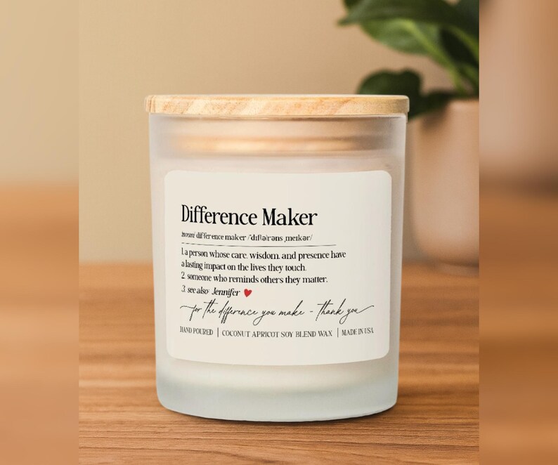 May include: A frosted glass candle with a wooden lid. The label reads "Difference Maker" with a definition and a handwritten message. The candle is hand-poured with coconut apricot soy blend wax.
