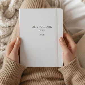 May include: A white notebook featuring the text "OLIVIA CLARK LCSW 2026" in black. The notebook is held by a person wearing a beige jumper. The notebook has a white elastic band.