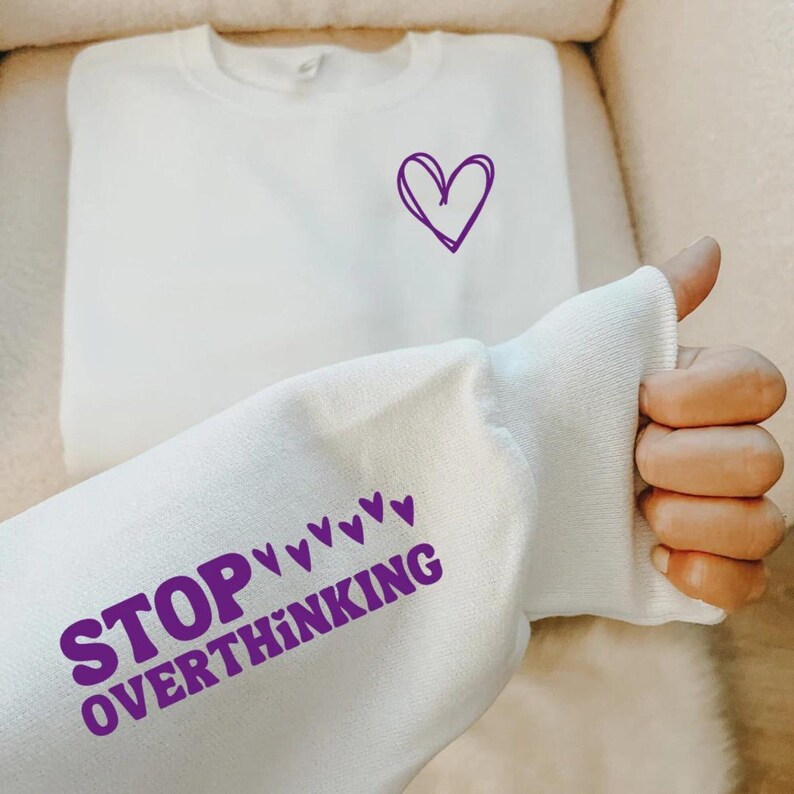 K&ouml;nnte beinhalten: Wei&szlig;er Pullover mit lilafarbenen Text "STOP overthinking" und einem kleinen lilafarbenen Herz auf der Brust.