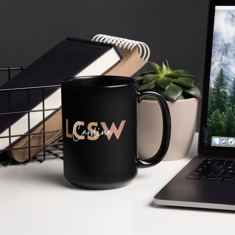 以下が含まれることがあります： 白い文字で「LCSW Caroline」とスタイリッシュなフォントで書かれた黒いセラミック製のコーヒーカップ。 「LCSW」の文字は、ピーチとライトブラウンの色です。