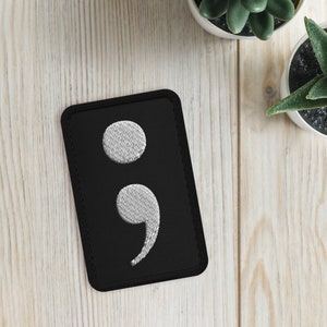 Könnte beinhalten: Schwarzer Stoff-Patch mit einem weißen gestickten Semikolon.