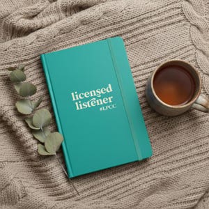 May include: A teal hardcover notebook with an elastic closure band, featuring the white text "licensed listener" and "#LPCC" on the front cover, accompanied by a sprig of eucalyptus and a ceramic mug filled with brown liquid.