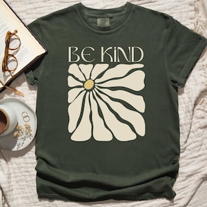 Pode incluir: Uma camiseta verde escura com uma margarida branca e o texto "BE KIND" estampado na frente.