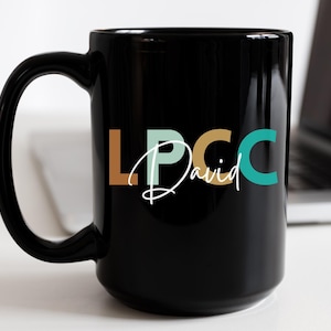 以下が含まれることがあります： 白い文字で「LPCC David」と書いてある黒いセラミック製のマグカップ。文字はオレンジ、グリーン、ターコイズブルーです。