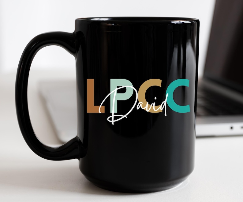 以下が含まれることがあります： 白い文字で「LPCC David」と書いてある黒いセラミック製のマグカップ。文字はオレンジ、グリーン、ターコイズブルーです。