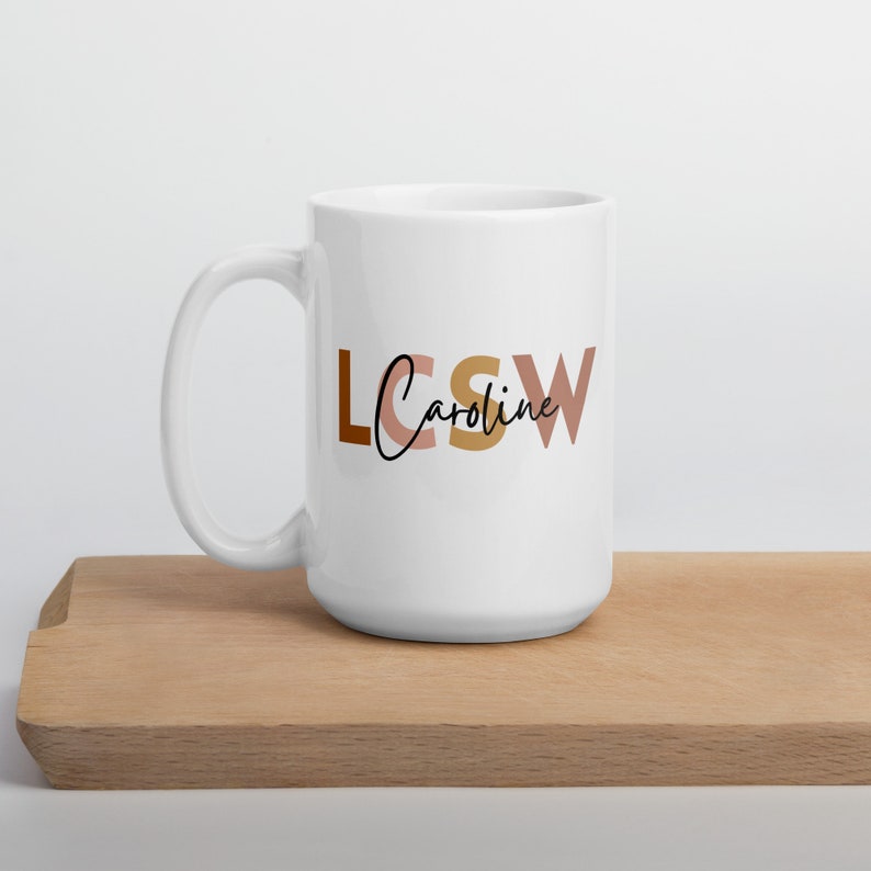 以下が含まれることがあります： ブラウン、ピンク、ゴールドの文字で「LCSW Caroline」と書かれた白いセラミックマグカップです。