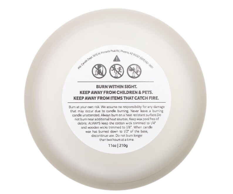 May include: A round, off-white candle with a white label. The label features safety warnings: "BURN WITHIN SIGHT," "KEEP AWAY FROM CHILDREN & PETS," and "KEEP AWAY FROM ITEMS THAT CATCH FIRE." The candle weighs 11oz.