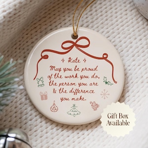 Op de afbeelding: Een rond wit keramieken ornament met een gouden ophangkoord. Het heeft een rode lintafbeelding aan de bovenkant en de tekst: Kate, May you be proud of the work you do, the person you are & the difference you make. Kleine vakantie-iconen omringen de tekst.