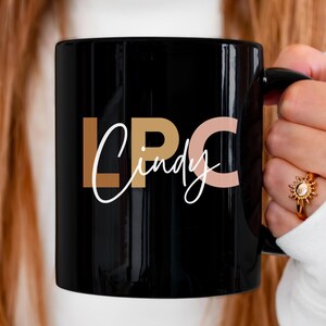 以下が含まれることがあります： 黒色のセラミック製のマグカップに、金とピンクの文字で「LPC Cindy」と書いてあります。