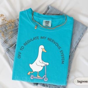 May include: A turquoise t-shirt featuring a cartoon goose on a scooter, with the text "OFF TO REGULATE MY NERVOUS SYSTEM". The shirt is folded on top of blue jeans and a gold necklace. The shirt is a Comfort Colors brand.