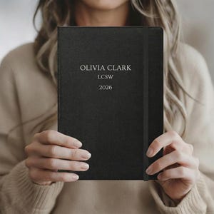 Puede incluir: Un diario negro con el nombre "OLIVIA CLARK", las letras "LCSW" y el año "2026" impresos en blanco. El diario tiene un cierre de banda elástica negra.