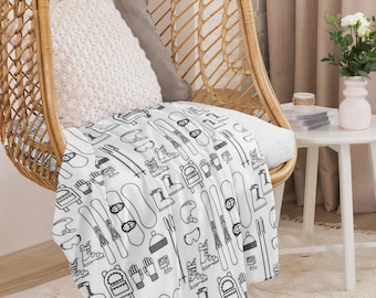 May include: A white throw blanket with a black and white pattern of winter sports equipment, including skis, snowboards, boots, and gloves. The blanket is draped over a wicker chair.