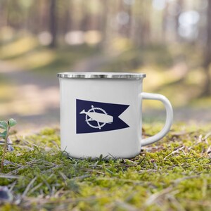 May include: White enamel camping mug with a blue and white nautical flag design featuring a whale and a rope wheel. The mug has a silver rim.